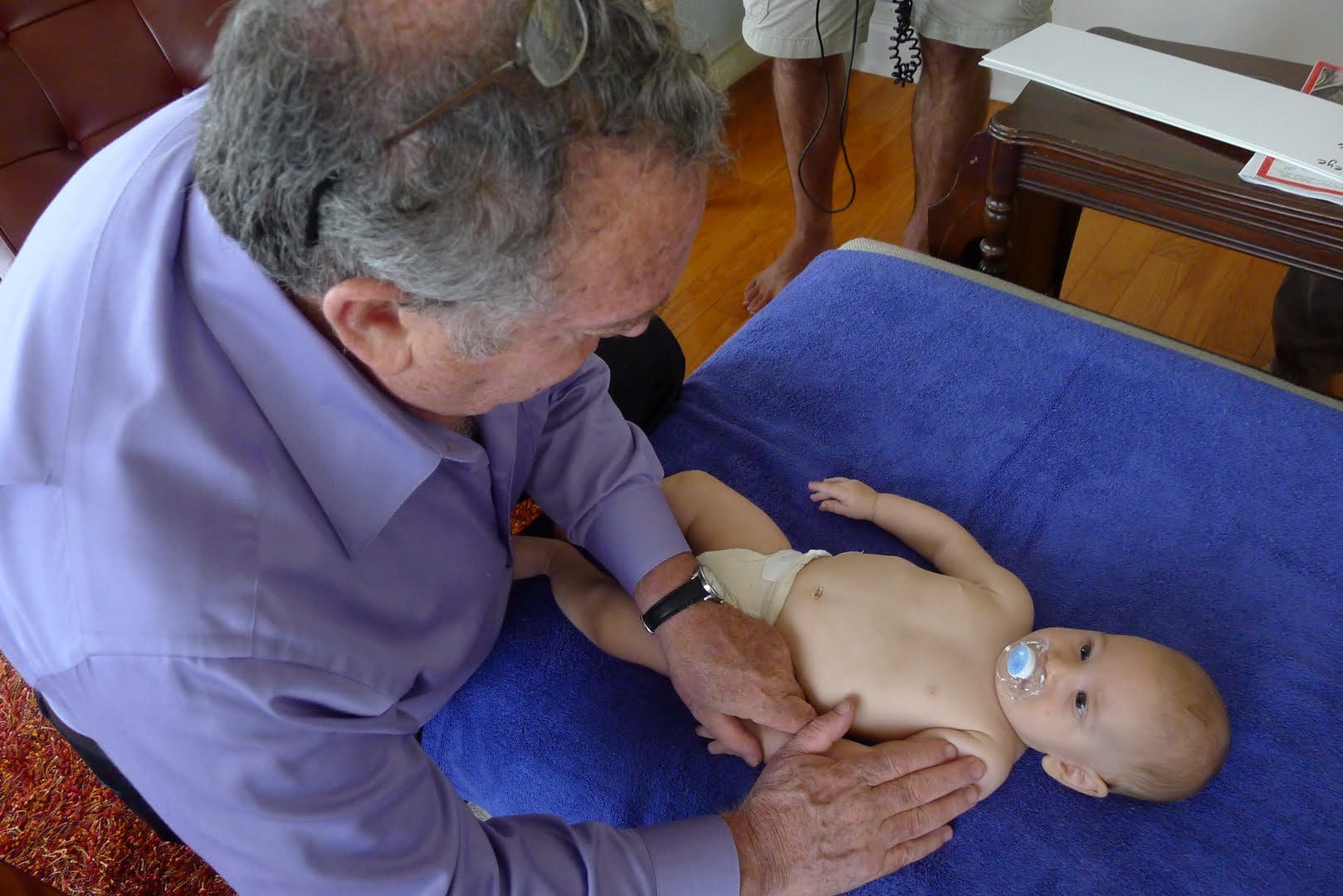 An older man wearing glasses and a purple shirt gently examines a baby with a pacifier lying on a blue towel-covered table.