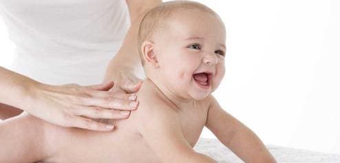 An adult gives a smiling baby a gentle back massage on a white surface.