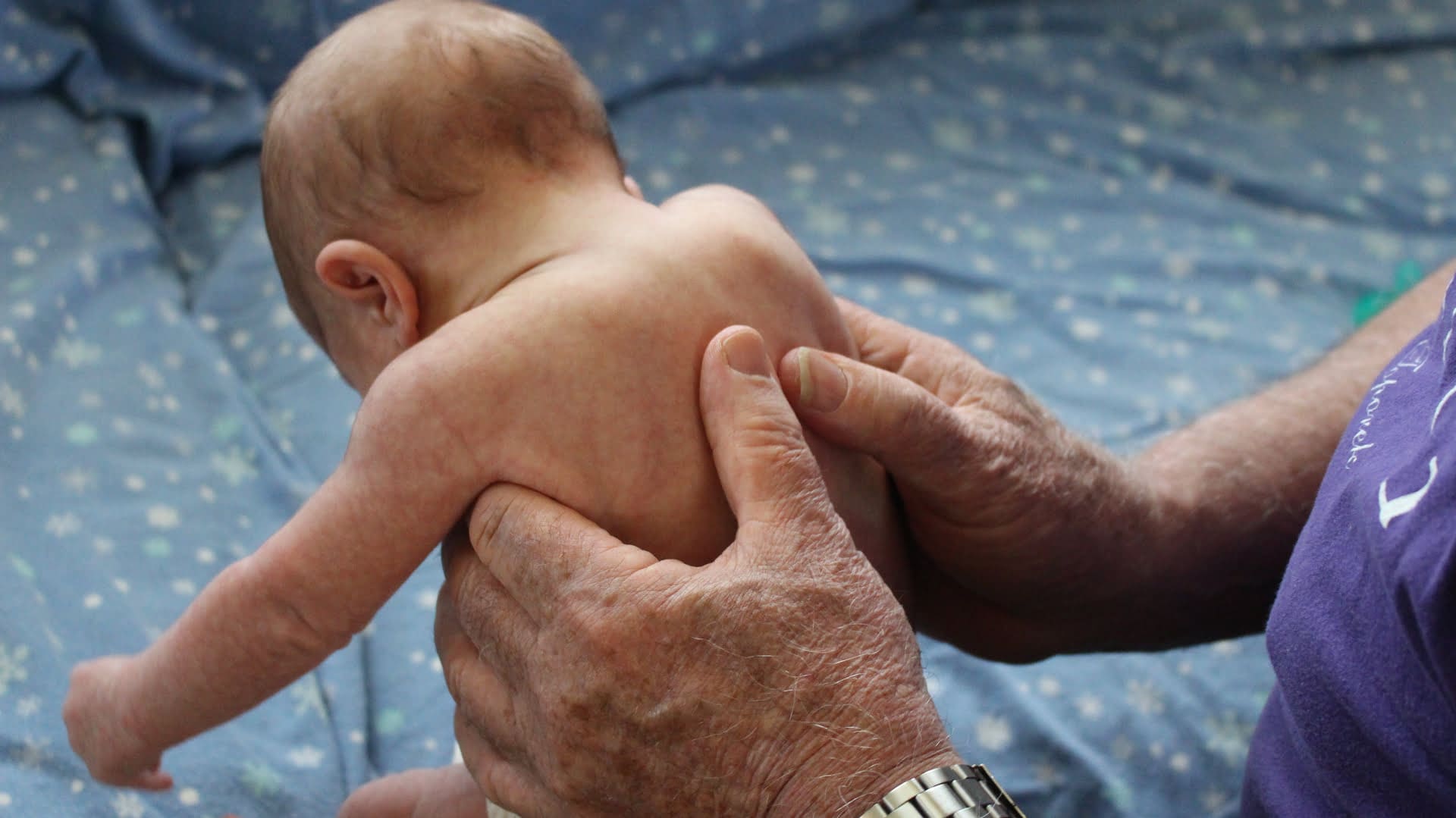 An adult with a watch holds a baby who is resting on their lap, on top of a light blue blanket with small white patterns.