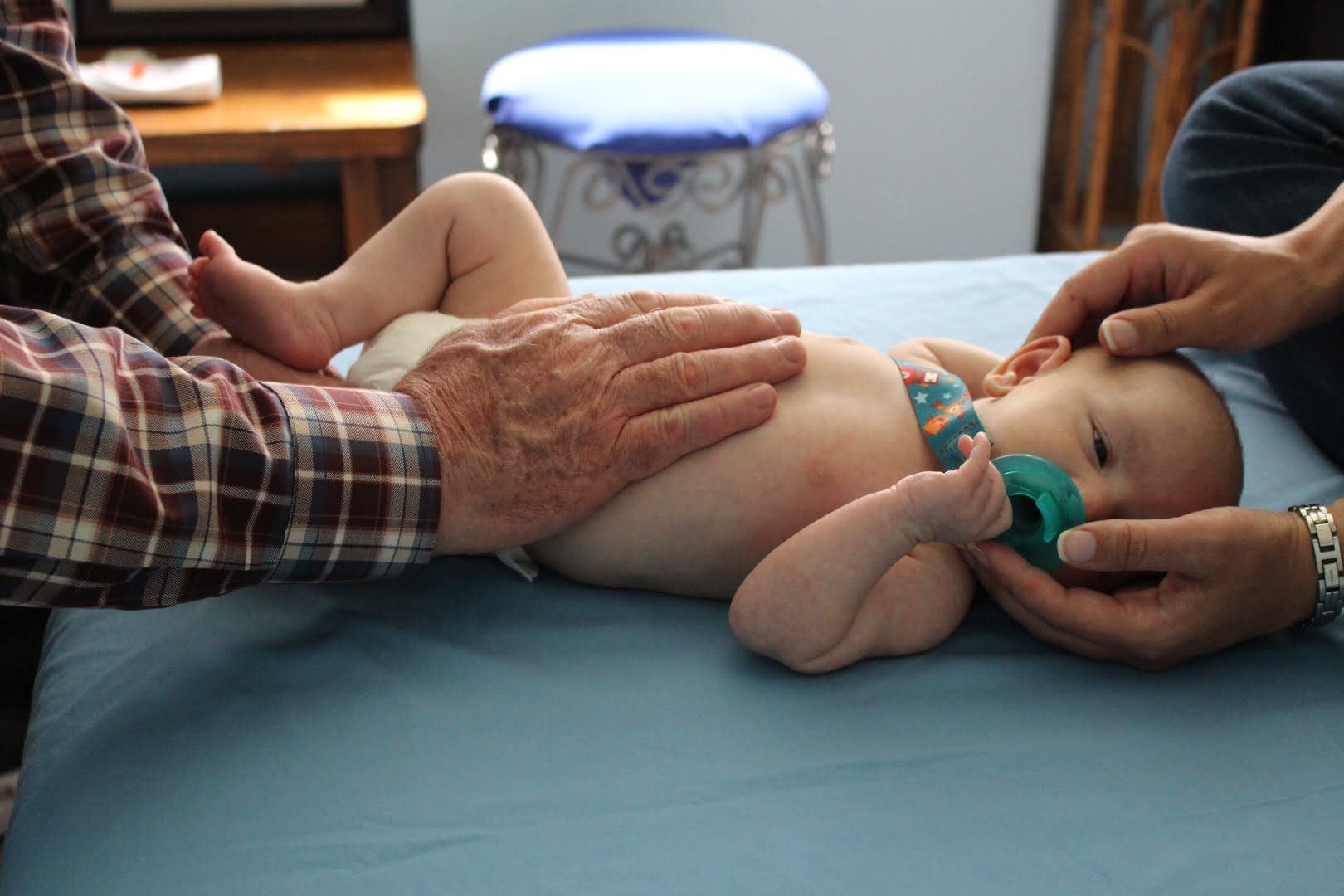 An infant lies on a blue surface with a pacifier in its mouth while being gently held by two adults wearing plaid and striped shirts.