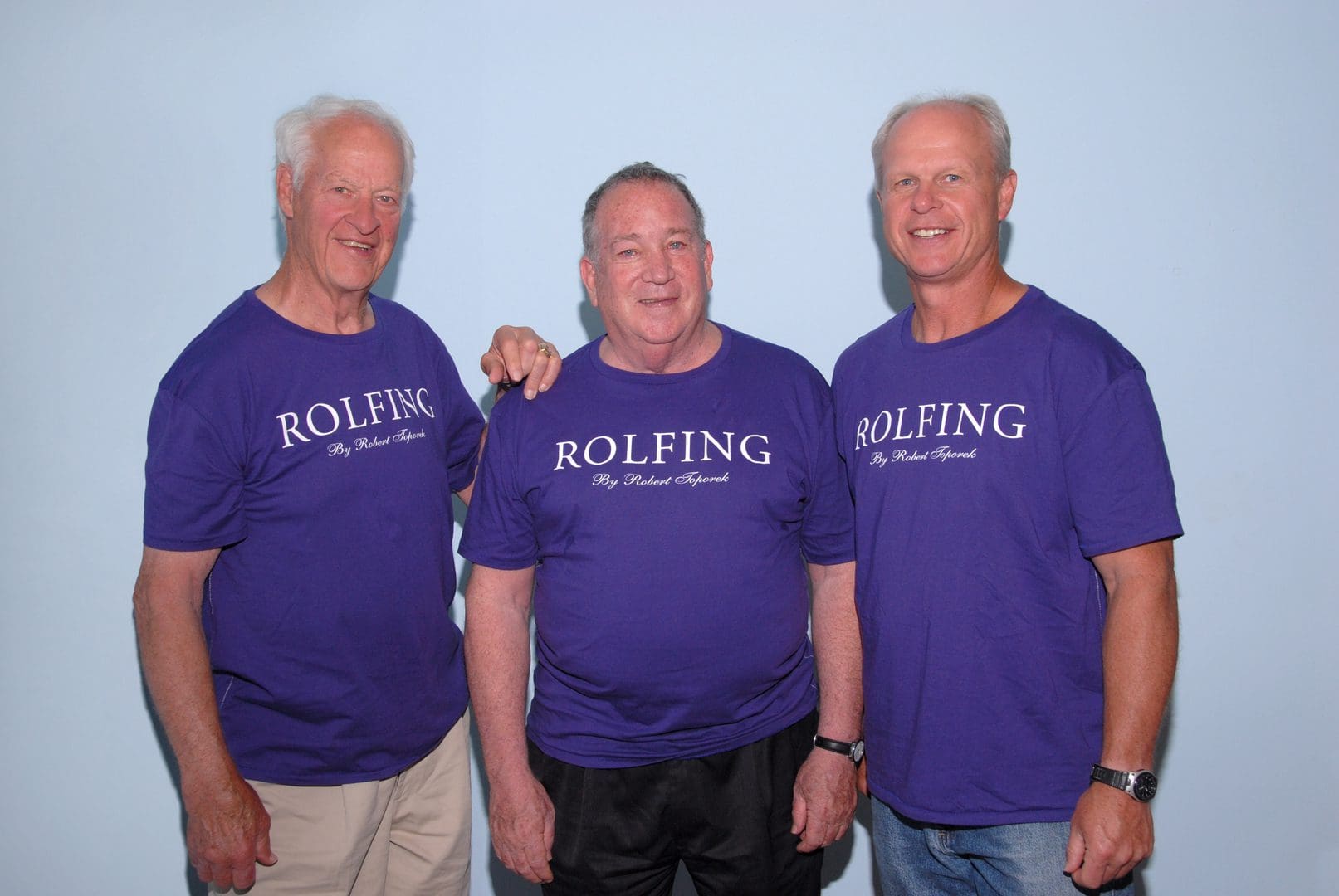 Three men wearing matching purple "ROLFING" t-shirts stand against a light blue backdrop, with the middle man placing his arm on the shoulder of the man on the left.