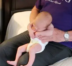 A baby wearing a white diaper sits on an adult's lap, bending forward with the adult's hands supporting the baby. The adult wears a purple shirt and a wristwatch.