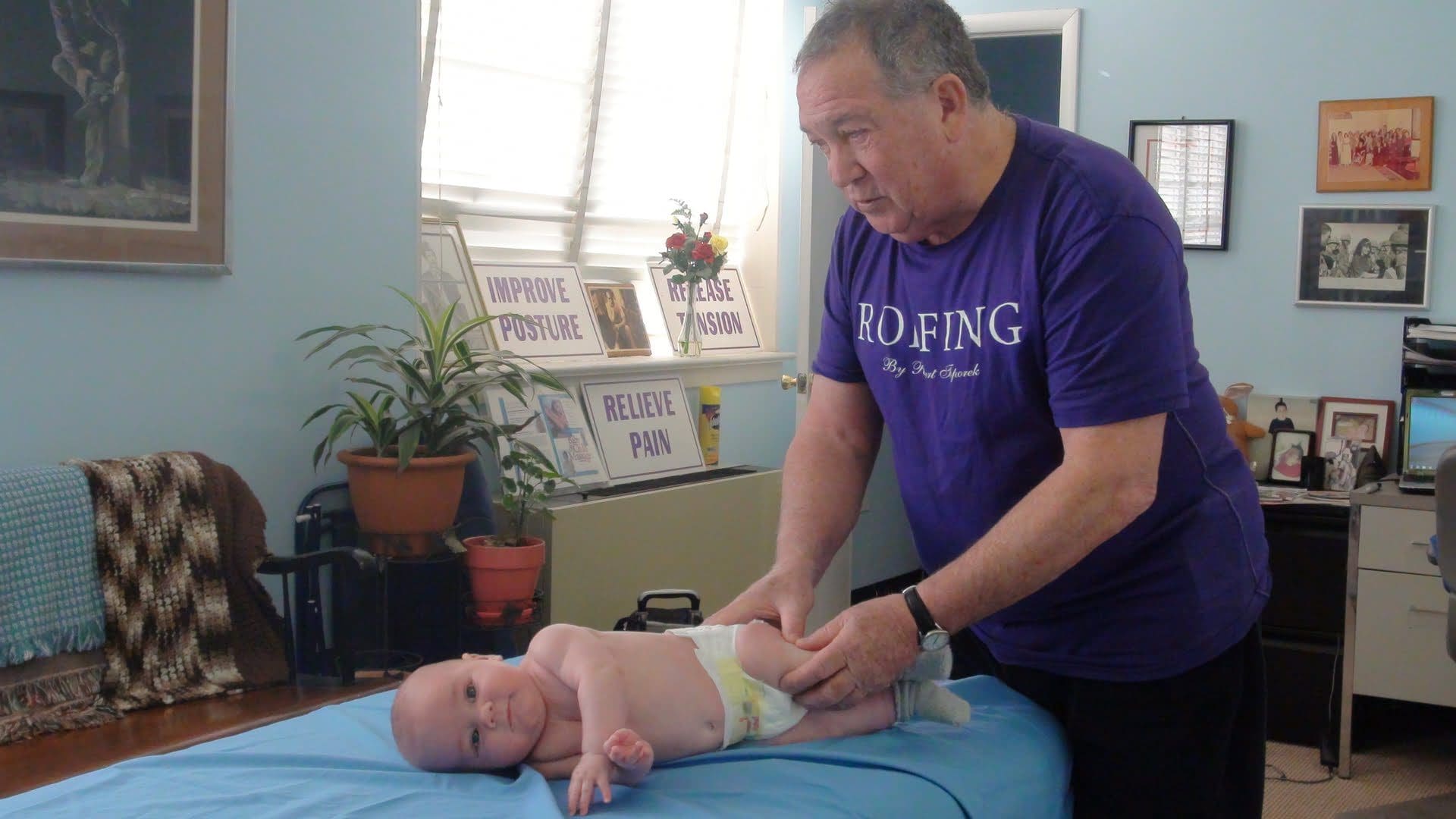 An adult is massaging an infant lying on a table in a room with various framed pictures and posters on the walls.