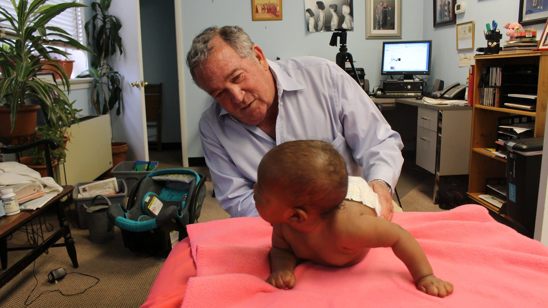 A man examines a baby who is laying on a pink blanket in an office setting with plants, a computer, and various items in the background.