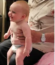 A smiling baby in a diaper is being held up by an older person wearing a floral shirt and watch.