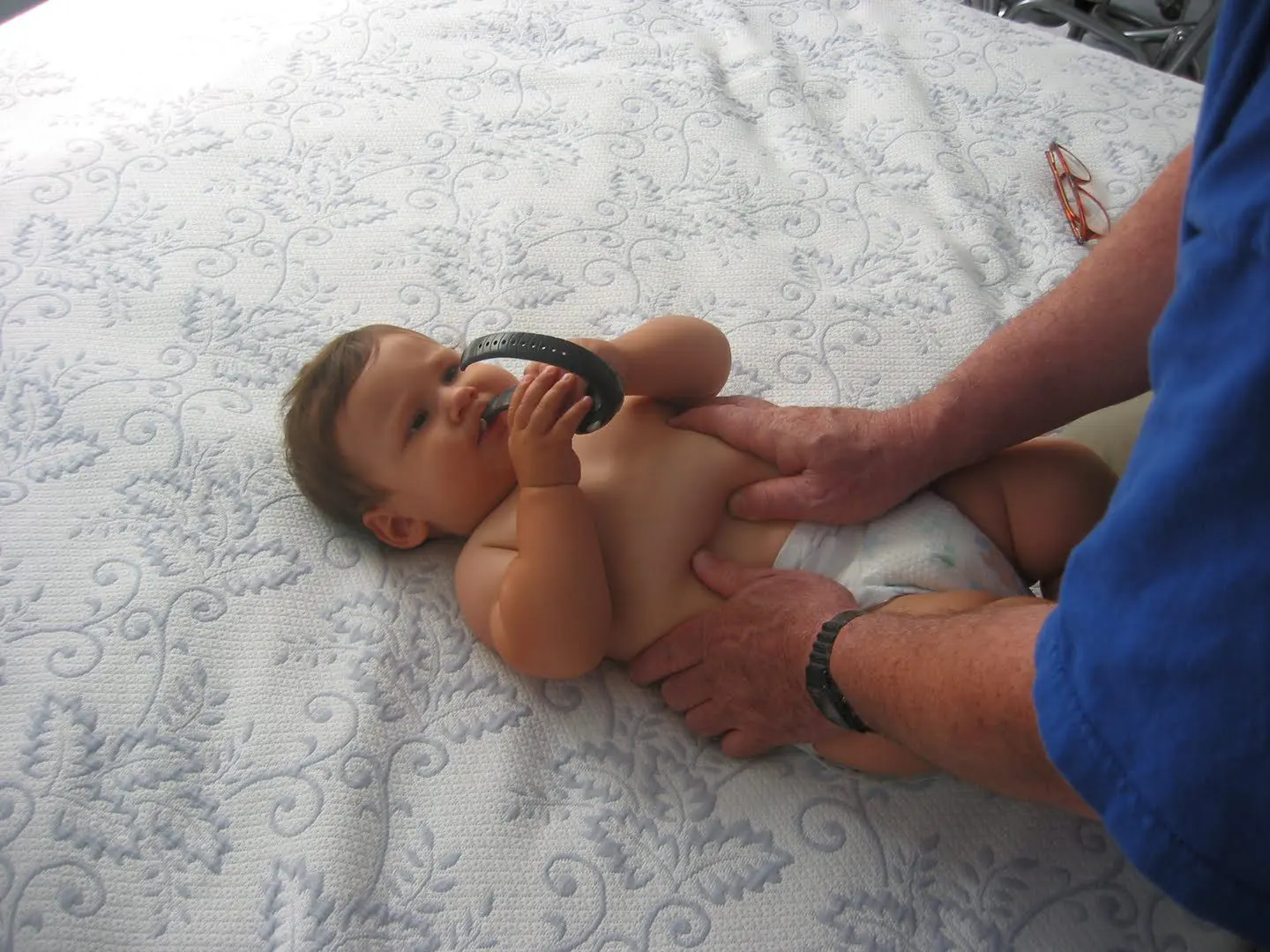 A baby lying on a patterned blanket is holding a teething toy while an adult's hands gently support the baby's torso.