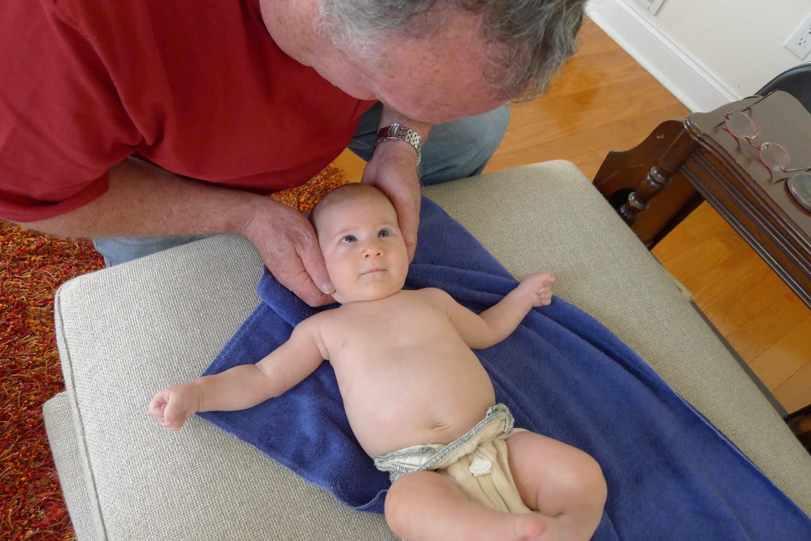 An older person in a red shirt gently holds a baby lying on a blue towel, who is looking up.