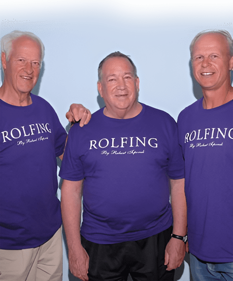 Three smiling men wearing matching purple "ROLFING" t-shirts, each with the text "Dr. Richard Apllund." The background is a plain light blue wall.