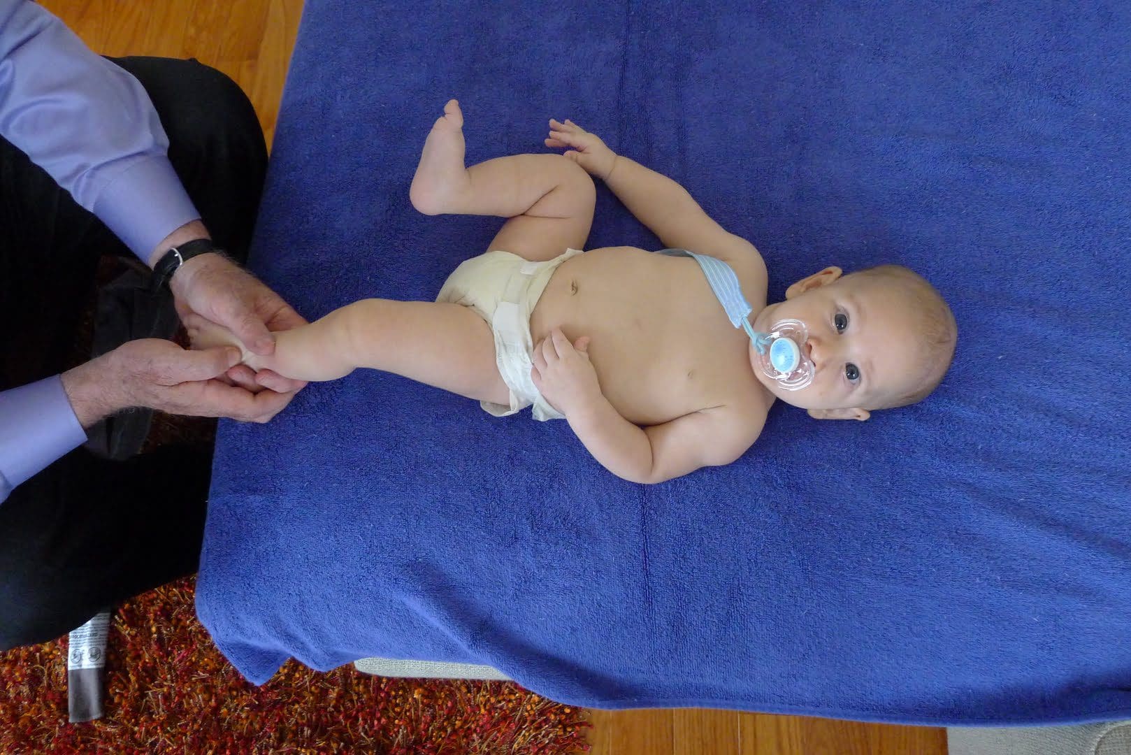 A baby wearing a diaper and pacifier lies on a blue blanket, while an adult gently holds one of the baby's legs.